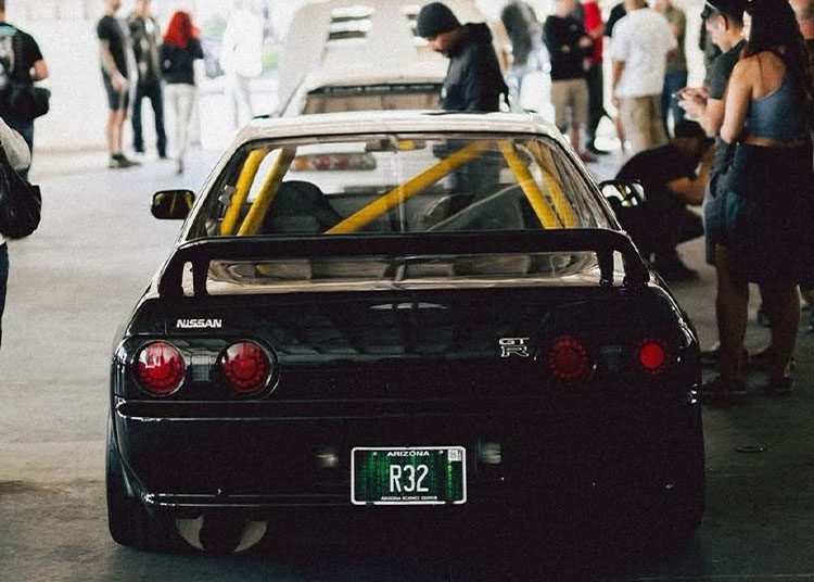 Rear view of a sleek black Nissan GT-R R32, proudly displaying its Arizona license plate at the duPont REGISTRY's Arizona Car Week 2025. Enthusiasts gather around, some capturing the moment on their phones as they admire this iconic masterpiece parked indoors.