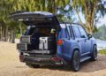 A blue Lexus Surf LX Concept SUV is parked near trees with its rear hatch open. Inside the trunk are a surfboard and camping gear, including a portable fridge and storage containers, perfect for adventure in Hawaii.