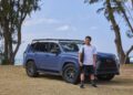 A person in a white shirt and black shorts stands beside a blue Lexus SUV on a grassy area near the ocean, with trees in the background.