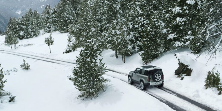 A silver SUV navigates a narrow, snow-covered road amid dense pine trees and majestic mountains, braving snowstorm conditions with determination.