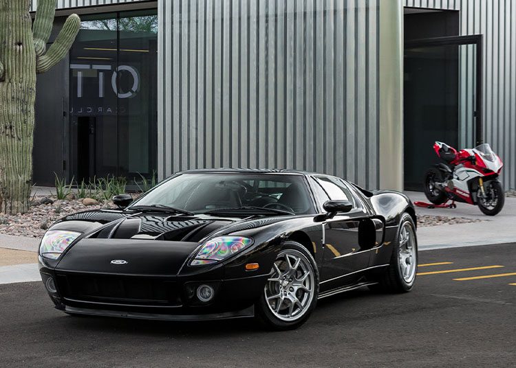 A sleek black sports car is parked in front of a modern building, with a motorbike peeking out in the background. This gem looks ready for the spotlight at Arizona Car Week 2025, where enthusiasts and collectors alike flock to events often highlighted in the duPont REGISTRY.