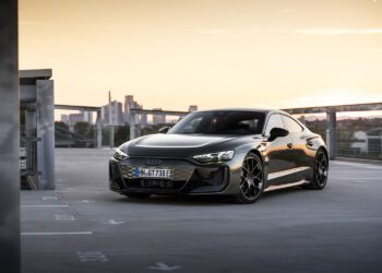 A sleek, black sports car is parked on a rooftop with a cityscape and sunset in the background.