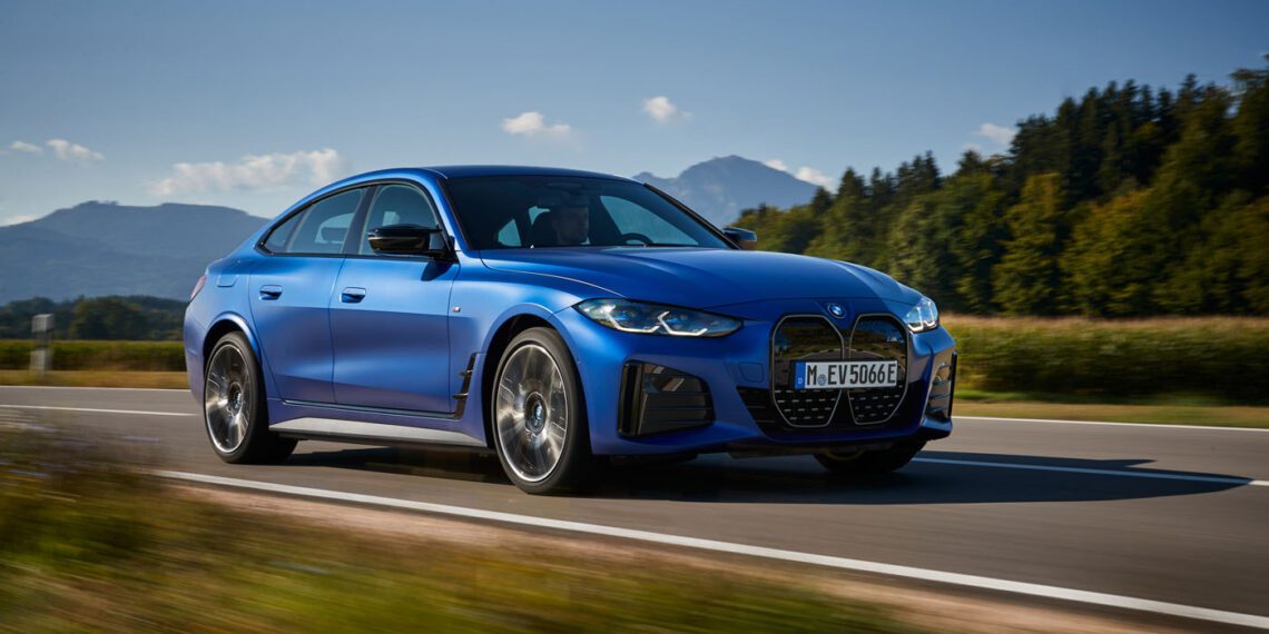 A 2024 BMW M blue sedan cruises on a rural road flanked by fields and trees, with majestic mountains in the background under a clear sky, embodying its record sales success.