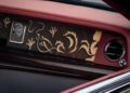 A close-up of the bespoke dashboard in a Rolls-Royce Phantom Dragon showcases a wooden panel with intricate golden dragon designs, complemented by a clock and round air vent, all elegantly framed by rich red leather trim.