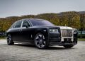 A bespoke black luxury car, reminiscent of a Rolls-Royce Phantom, is parked on a cobblestone surface. Trimmed trees and a stately building stand in the background beneath the cloudy sky.