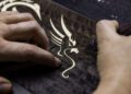 A person skillfully carves an intricate Phantom Dragon design onto a dark, textured surface using a carving tool, creating a bespoke masterpiece.