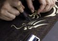 Hands meticulously carving an intricate Phantom Dragon design into wood using a sharp tool, embodying the bespoke craftsmanship akin to a Rolls-Royce.