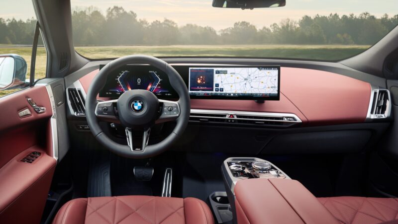 Interior of a car featuring a digital dashboard, steering wheel with logo, and red leather seats, set against a background of trees and open fields.