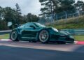 A sleek, dark green Porsche 911 GT3 RS speeds around a race track, surrounded by trees and a metal fence.