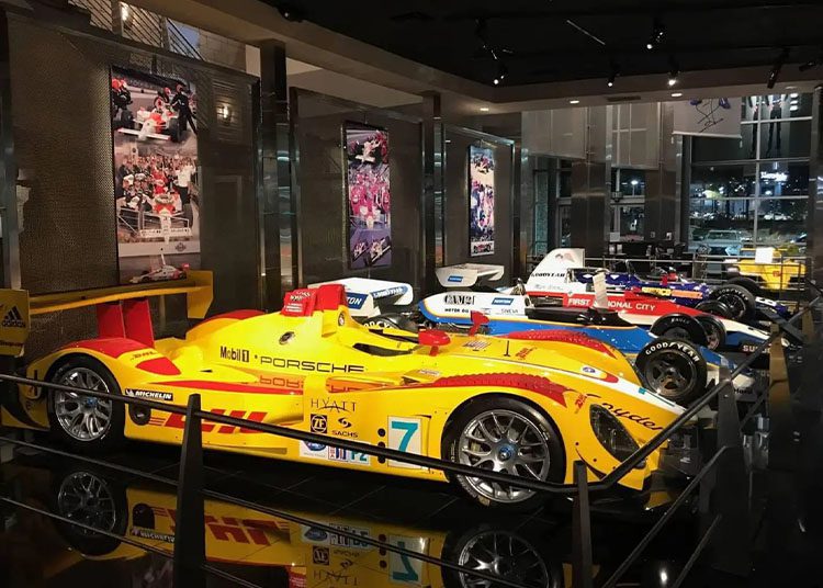 Several racing cars, highlighted in the duPont REGISTRY, are displayed in a museum exhibit. They’re positioned in a row with informational panels and posters in the background, creating an engaging tribute just in time for Arizona Car Week 2025.