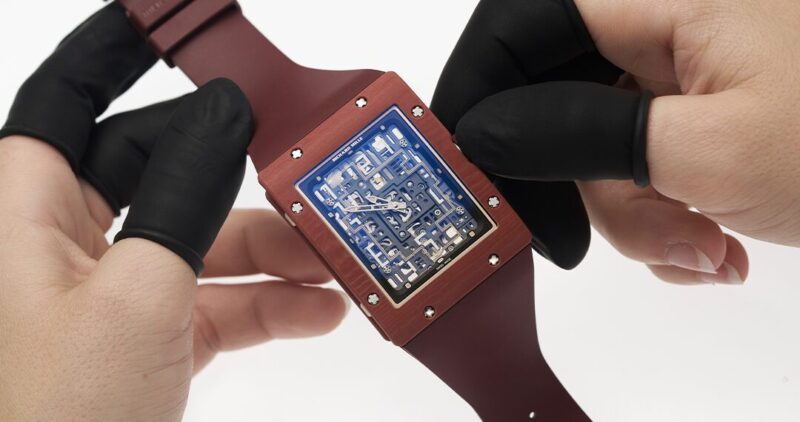 Hands with gloves delicately hold a luxury wristwatch, featuring a striking blue geometric face, set against its vibrant red band.