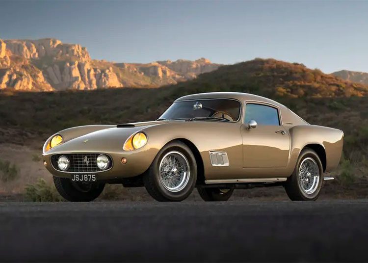 A classic gold sports car, shimmering in the sunset, sits parked on a road with majestic mountains in the background. This stunning scene could grace the pages of duPont REGISTRY or be a highlight at Arizona Car Week 2025.