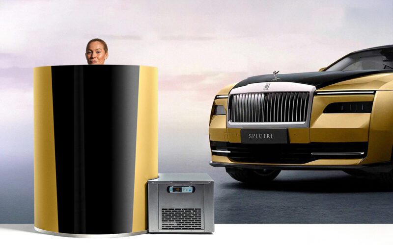 A person standing in a yellow and black cylindrical cryotherapy chamber next to a luxury car with a "Spectre" license plate.