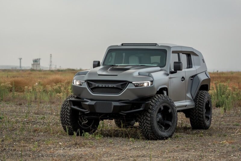 A unique creation, this matte gray armored SUV with large off-road tires sits majestically on a gravel landscape under an overcast sky, reflecting the pinnacle of custom car craftsmanship.