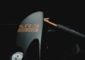 Close-up of a sleek black object with orange text "SR3 XXR 3000." As Radical Motorsports celebrates its 28th Anniversary, visible details include a metal fuel cap and a black handlebar marked "RAD." The dark scene is highlighted with focused lighting.