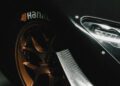 Close-up of a Radical Motorsports sports car wheel with bronze alloy rims, showcasing a prominent tire brand logo alongside a sleek carbon fiber body panel. The headlight is partially visible against the black background, celebrating innovation in every detail on this special anniversary edition.