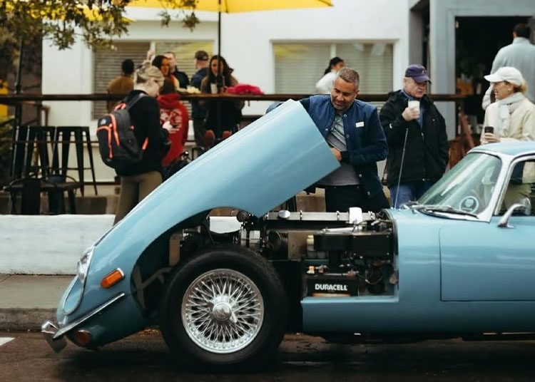 During Arizona Car Week 2025, a man examines the open hood of a vintage blue car parked on a lively street, with curious onlookers and a bustling café in the background.