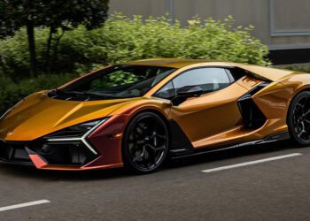 A sleek, Oro Alba-colored sports car with black accents drives on a road, surrounded by greenery. This stunning Revuelto model turns heads as it reflects the style and sophistication associated with Lamborghini Dallas.
