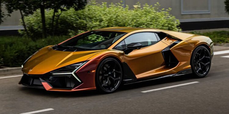 A sleek, Oro Alba-colored sports car with black accents drives on a road, surrounded by greenery. This stunning Revuelto model turns heads as it reflects the style and sophistication associated with Lamborghini Dallas.