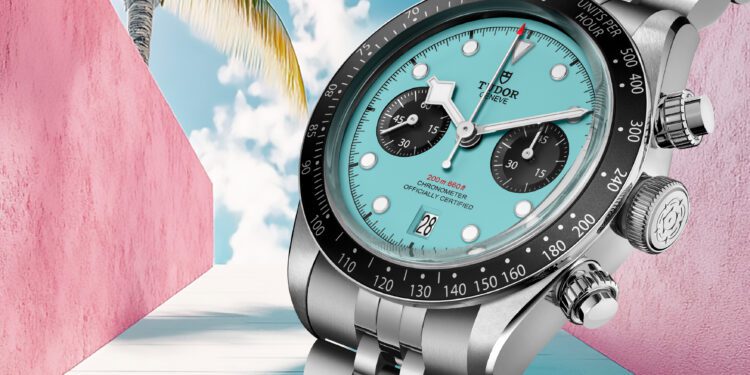 A TUDOR Black Bay Chrono wristwatch with a dazzling turquoise dial and black subdials is elegantly displayed against a pink-walled alley, where a lone palm tree sways under the Flamingo Blue sky.