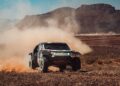 A Dacia rally car speeds through the dusty desert landscape of the Dakar Rally, with mountains looming in the background.