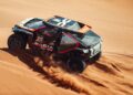 A race car from the Dakar Rally navigates through a sandy desert terrain, kicking up a trail of dust.