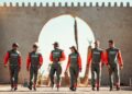 Six Sandriders in matching racing suits stride confidently together outdoors, set against the backdrop of a large, arched structure—a scene reminiscent of the legendary Dakar Rally.