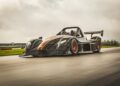 A sleek, low-profile black race car with orange accents, marking Radical Motorsports' 28th Anniversary, speeds around the racetrack on a cloudy day.