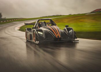 A black race car with orange accents from Radical Motorsports speeds around a wet, curving track under an overcast sky, celebrating the company's 28th Anniversary.