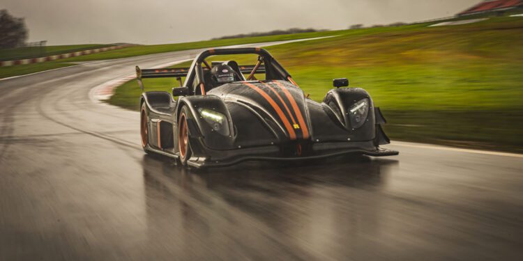 A black race car with orange accents from Radical Motorsports speeds around a wet, curving track under an overcast sky, celebrating the company's 28th Anniversary.