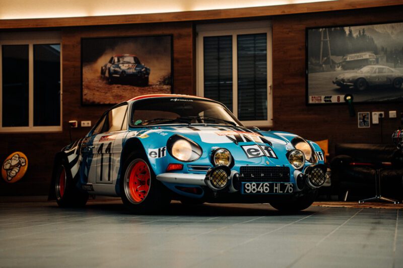 A vintage blue and white rally car with red rims is parked indoors, embodying the spirit of an Alpine Dream. The car features racing decals, evoking a sense of adventure. Framed photos and wood-paneled walls provide a nostalgic backdrop worthy of Film Friday.