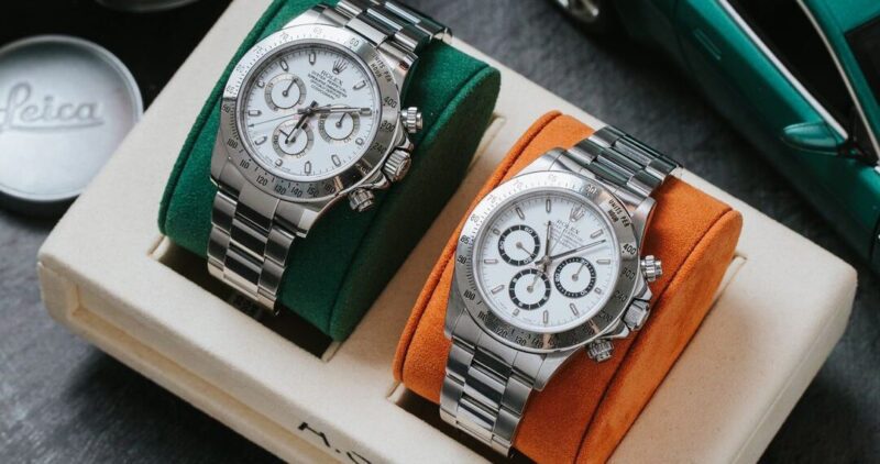 Two luxury stainless steel chronograph watches with white dials and black subdials, elegantly displayed on green and orange cushions. They sit near a car model and a Leica lens cap, embodying a sophisticated lifestyle aesthetic.