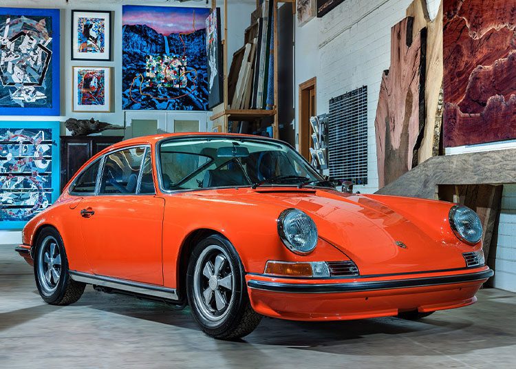 A vintage orange sports car, reminiscent of those featured in duPont REGISTRY, is parked indoors, surrounded by artworks and wooden materials. It feels like stepping into an exclusive gallery event at Arizona Car Week 2025.