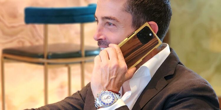 A man in a suit holds a large smartphone to his ear, its elegance elevated by an EXOvault Case. He sits indoors next to a chair, the craftsmanship of his attire matched by his watch.