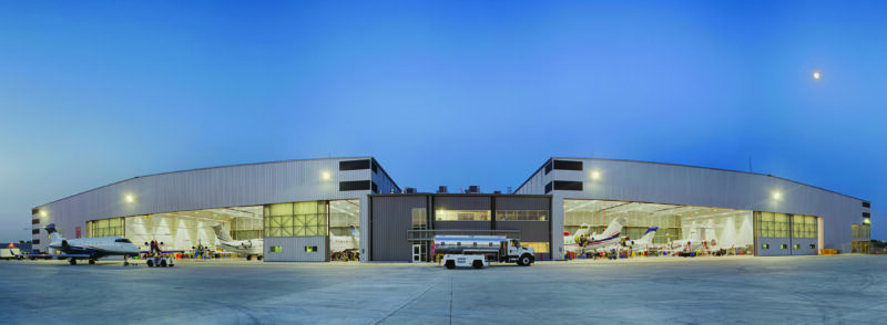 Panoramic view of a large aircraft hangar at twilight, featured in the duPont REGISTRY 2025 Aviation Guide, with several planes inside and ground support vehicles parked outside.