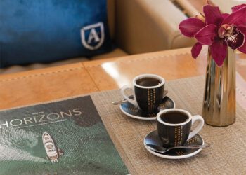 On a table next to a blue pillow, coffee cups sit alongside a book titled "Horizons" and a vase with purple orchids, evoking the elegance of private aviation.