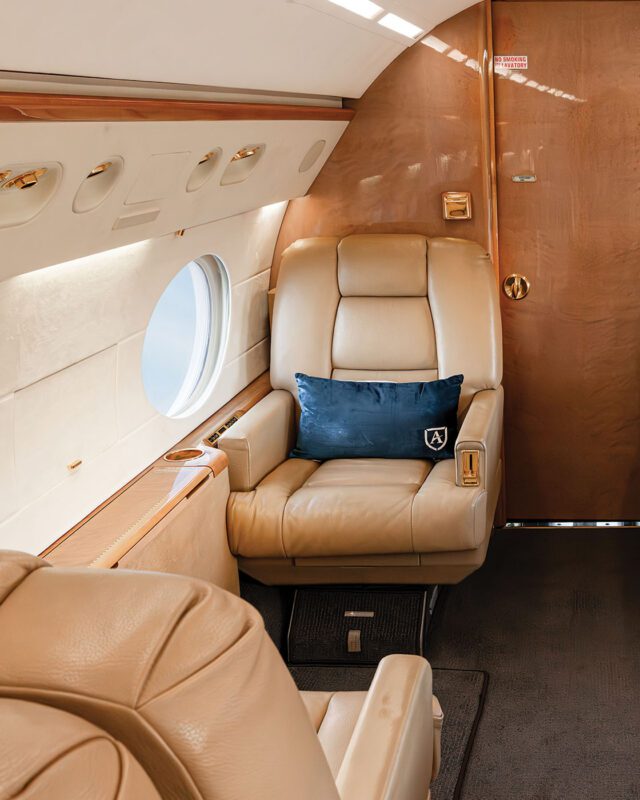 Luxurious Fly Alliance private jet interior with beige leather seats, a window view, elegant wooden paneling, and a blue decorative pillow on one seat. Soars above expectations in comfort and style.