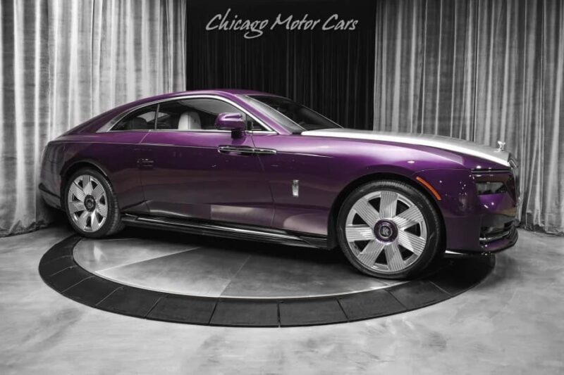 A purple and silver Rolls-Royce Spectre electric super coupe is displayed on a rotating platform inside a showroom with grey curtains and "Chicago Motor Cars" signage above.