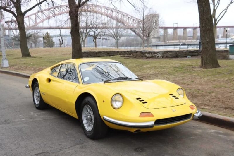 A yellow vintage Ferrari Dino is parked on a street next to a park, with trees and a bridge in the background.