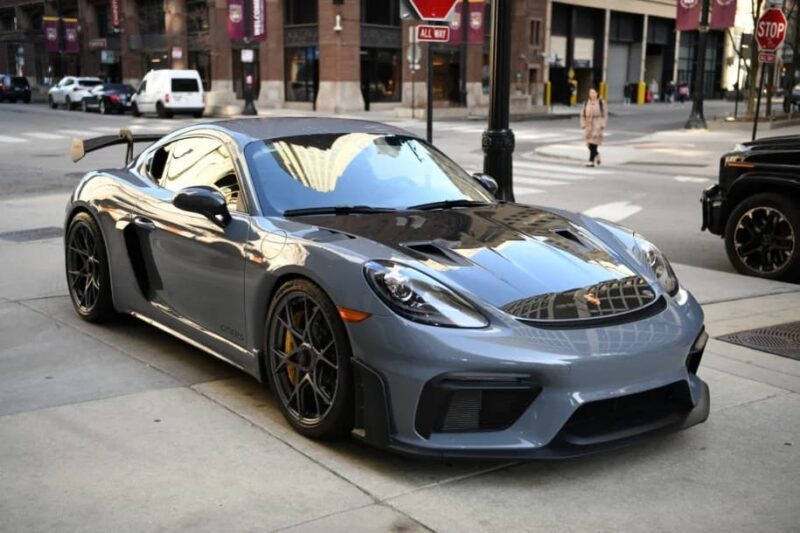 A gray Porsche 718 Cayman GT4 RS is parked on a city street near a crosswalk, showcasing Purist Performance. Buildings and a few pedestrians form the urban backdrop, perfect for those seeking high-end cars for sale.