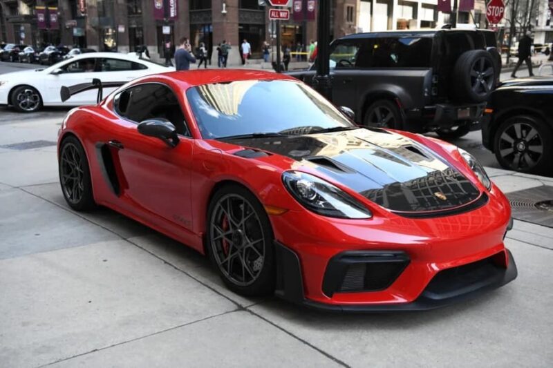 A red Porsche 718 Cayman GT4 RS with black accents is parked on a city street alongside other cars for sale, with buildings in the background.