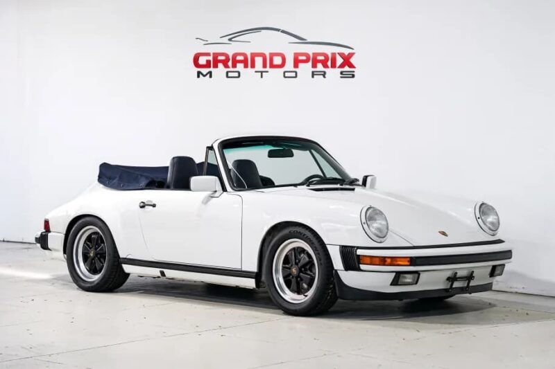A white Porsche 911 convertible with a dark soft top, known among the best manual transmission cars, is parked indoors in front of a white wall featuring the Grand Prix Motors logo.