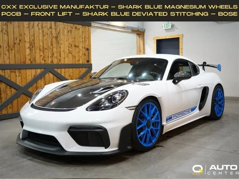 White Porsche 718 Cayman GT4 RS with black hood and shark blue wheels parked indoors, featuring Purist Performance decals, a carbon fiber front splitter, and sporty details—an eye-catching choice among cars for sale.