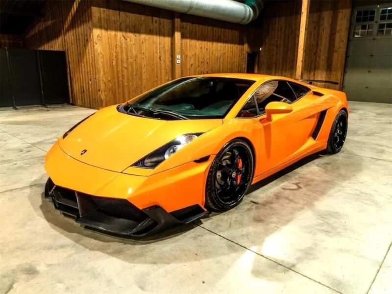 An orange Lamborghini sports car, one of the best manual transmission cars, is parked indoors on a concrete floor with wooden walls in the background.