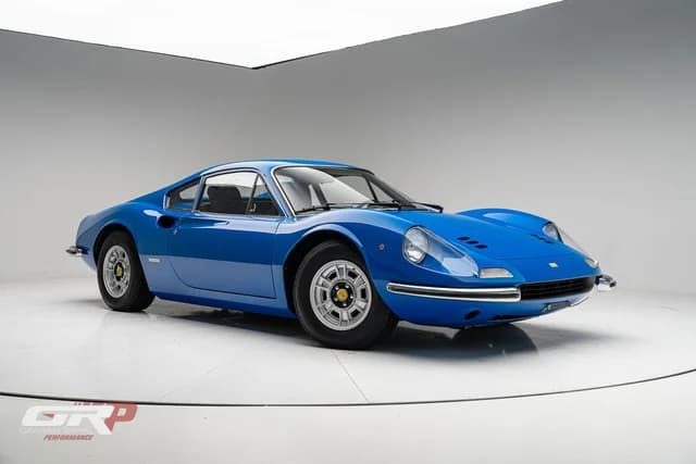 A blue classic Ferrari Dino is displayed in a white studio with minimal background, viewed from the front left side.