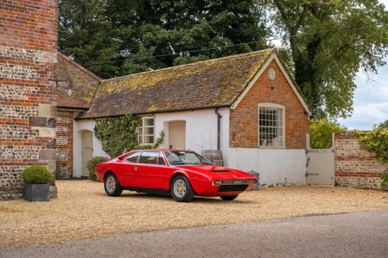 A red Ferrari Dino sports car is parked on a gravel driveway in front of a rustic brick and white cottage with a tiled roof, reflecting the timeless appeal of classic Ferrari cars.