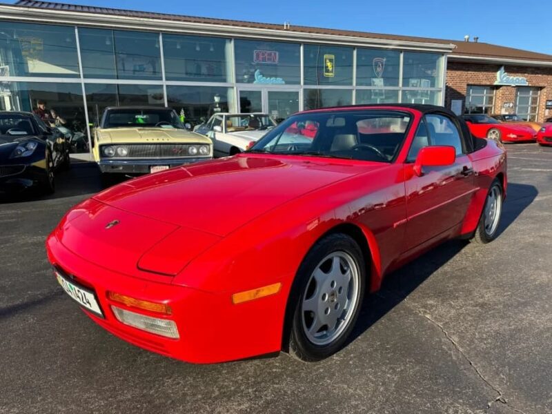 A red transaxle Porsche 944 convertible is parked outside a dealership on a sunny day, with several other cars in the background—a perfect chance to invest in one of the unsung classics.