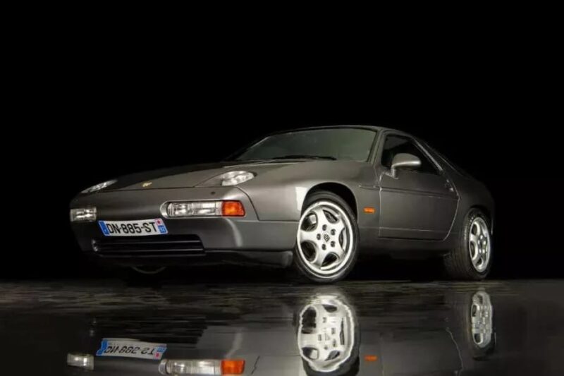 A dark grey Transaxle Porsche 928 sports car, one of the unsung classics worth a second look to invest in, is parked on a reflective surface against a black background, viewed from the front left.