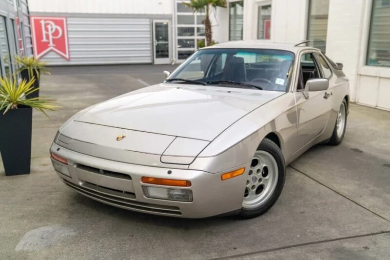 A silver Porsche 944, an unsung classic from the Transaxle Porsche lineup, is parked on a concrete surface in front of a modern building with glass windows and metal doors—a smart choice for those looking to invest.