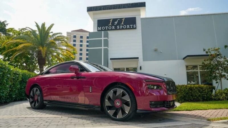 A maroon Rolls-Royce Spectre Electric Super Coupe is parked in front of a building with a sign reading "1 of 1 Motor Sports," surrounded by palm trees and greenery.